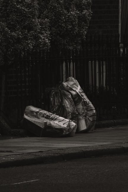 A large collection of mixed household waste and recyclable materials, including cardboard boxes, plastic bags, paper, and packaging, piled beside and overflowing from a grey communal rubbish bin marke